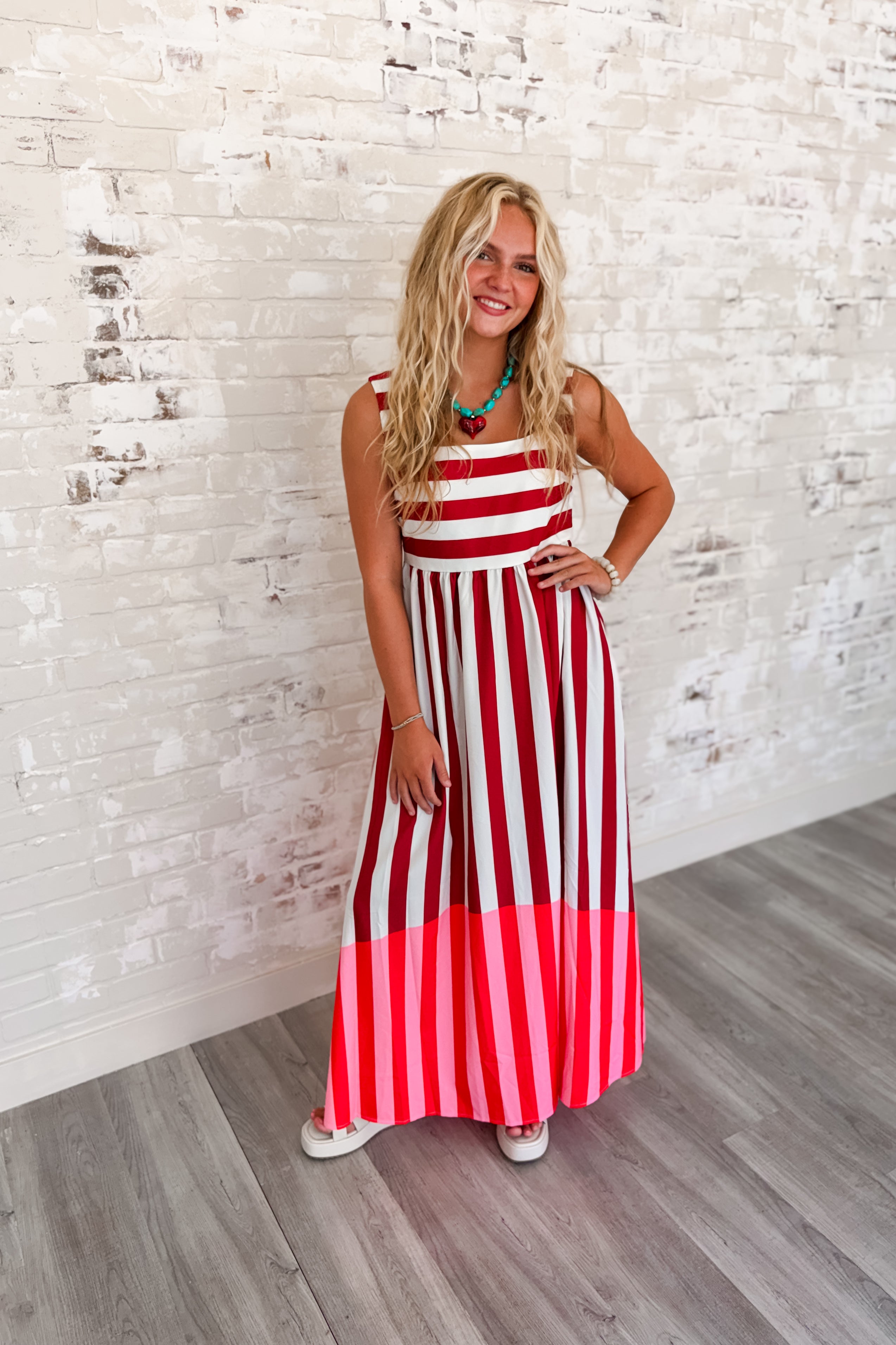 Woman wearing a sleeveless red and ivory striped maxi dress with a square neckline, flowing skirt, side pockets, and a vibrant pink color-block hem.