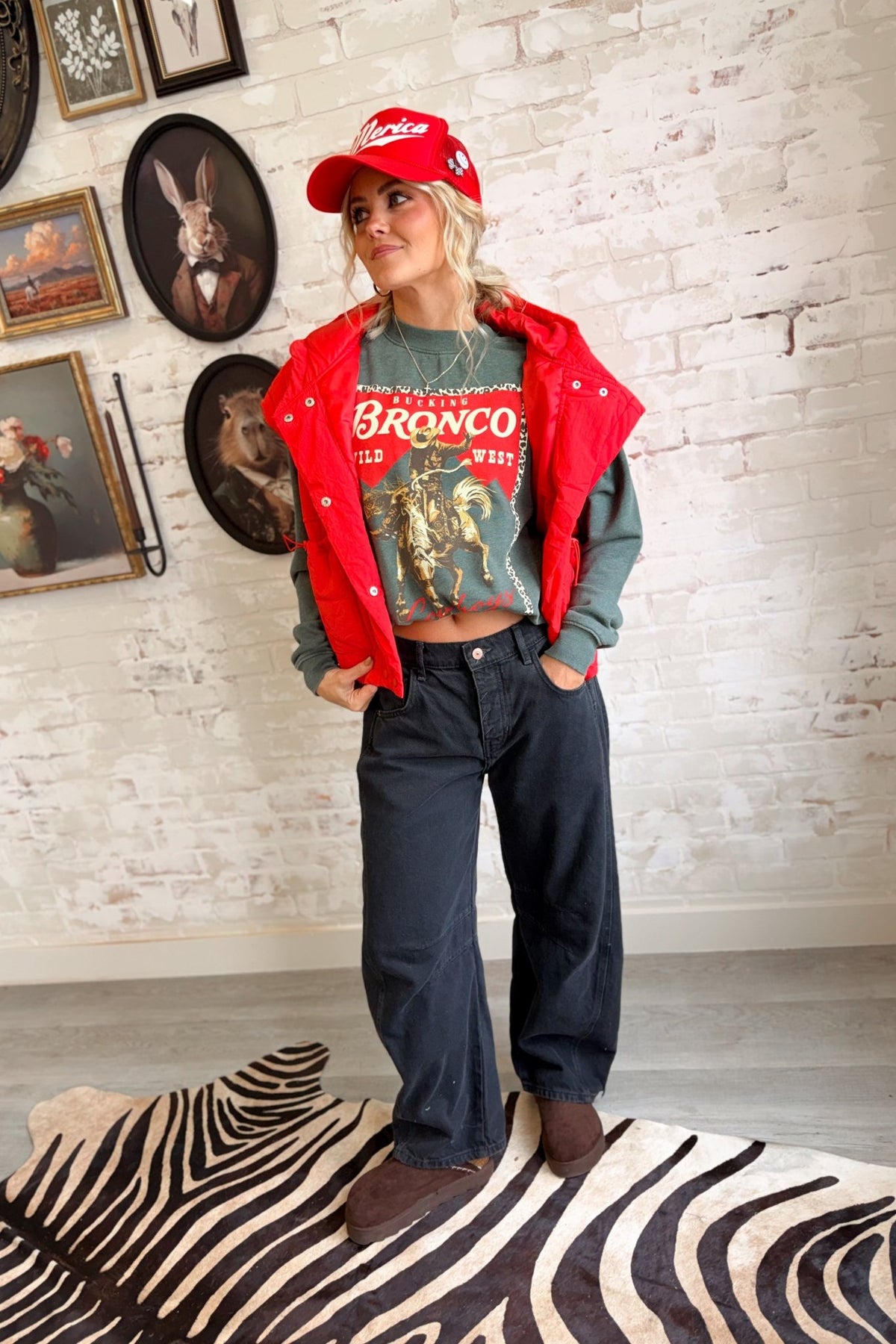 Women's vintage washed sage green western graphic crewneck sweatshirt featuring an oversized, relaxed silhouette, classic round neckline, dropped shoulders, long sleeves, and ribbed cuffs. The front showcases a bold retro rodeo-inspired graphic with a bucking bronco horse and cowboy rider in distressed golden yellow and brown tones, set against a vibrant red background with a leopard print border. Featuring the words "Bucking Bronco," "Wild West," and "Cowboys" in vintage western typography, this cozy pullo