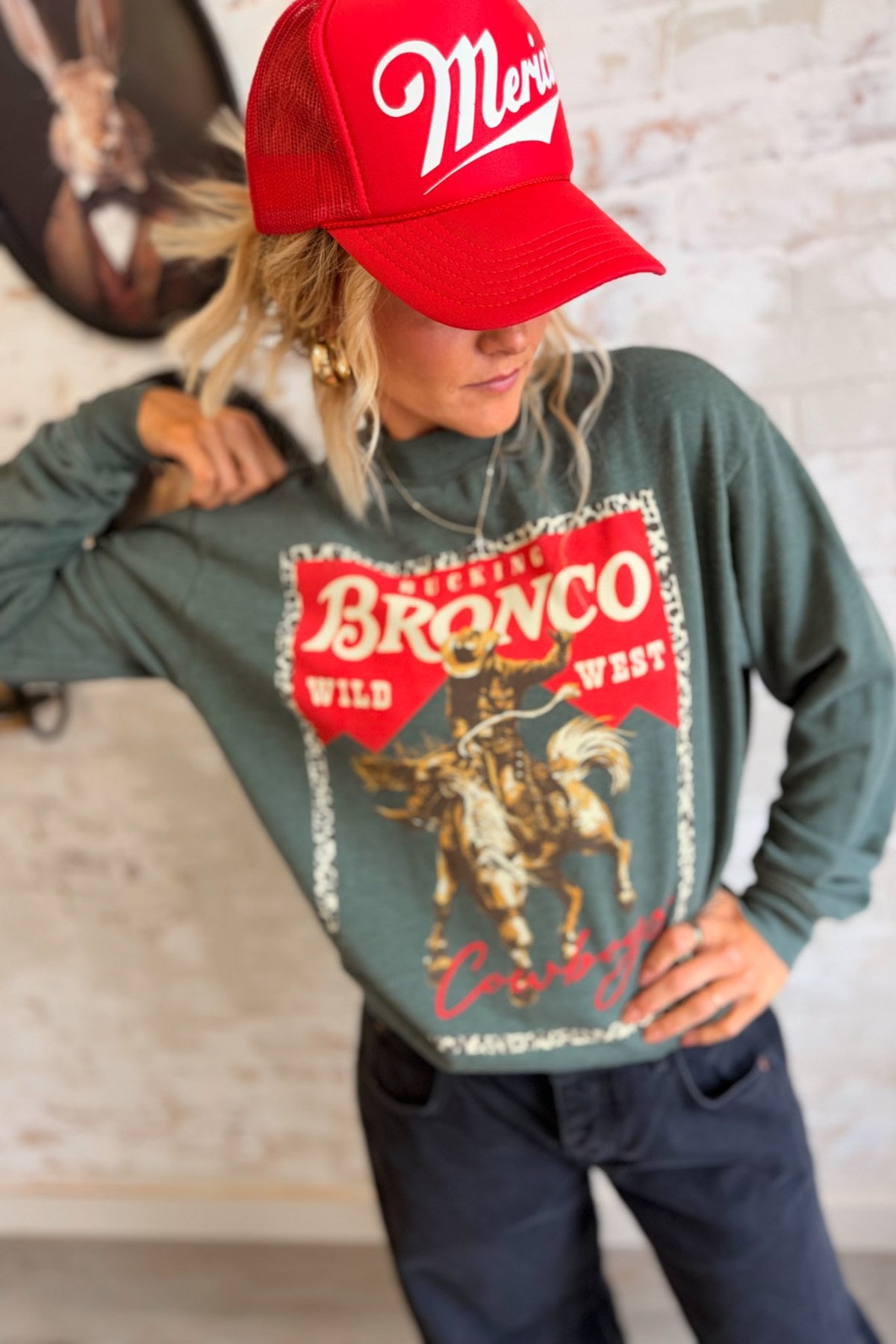 Women's vintage washed sage green western graphic crewneck sweatshirt featuring an oversized, relaxed silhouette, classic round neckline, dropped shoulders, long sleeves, and ribbed cuffs. The front showcases a bold retro rodeo-inspired graphic with a bucking bronco horse and cowboy rider in distressed golden yellow and brown tones, set against a vibrant red background with a leopard print border. Featuring the words "Bucking Bronco," "Wild West," and "Cowboys" in vintage western typography, this cozy pullo