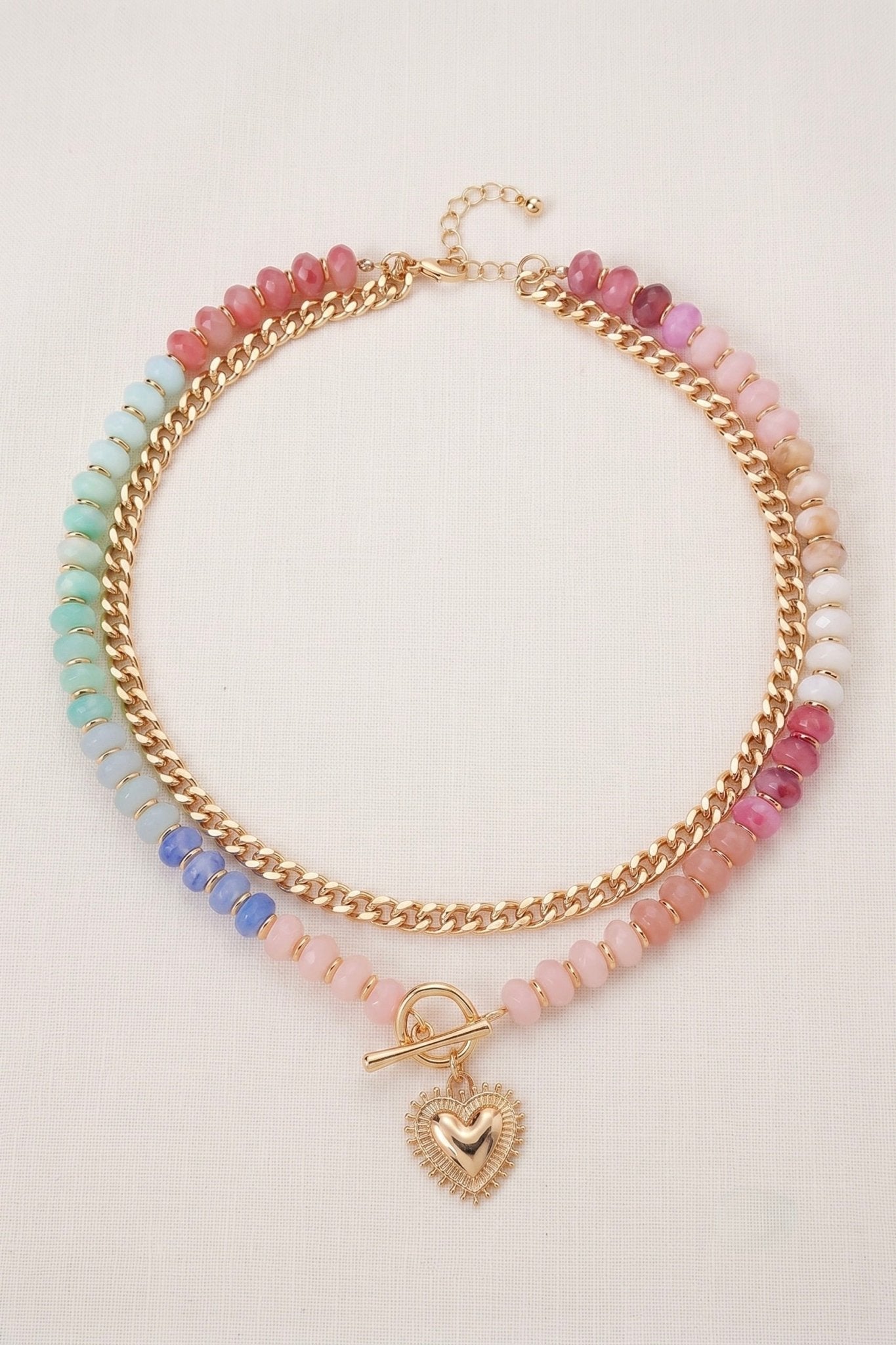 Layered pastel beaded necklace with gold chain detail, toggle clasp, and radiant heart charm pendant in pink, blue, peach, and green tones.