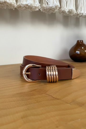 Close-up of a women's Brown faux leather belt with a shiny rose gold-toned buckle and five decorative rose gold rings, displayed coiled on a light wooden surface.