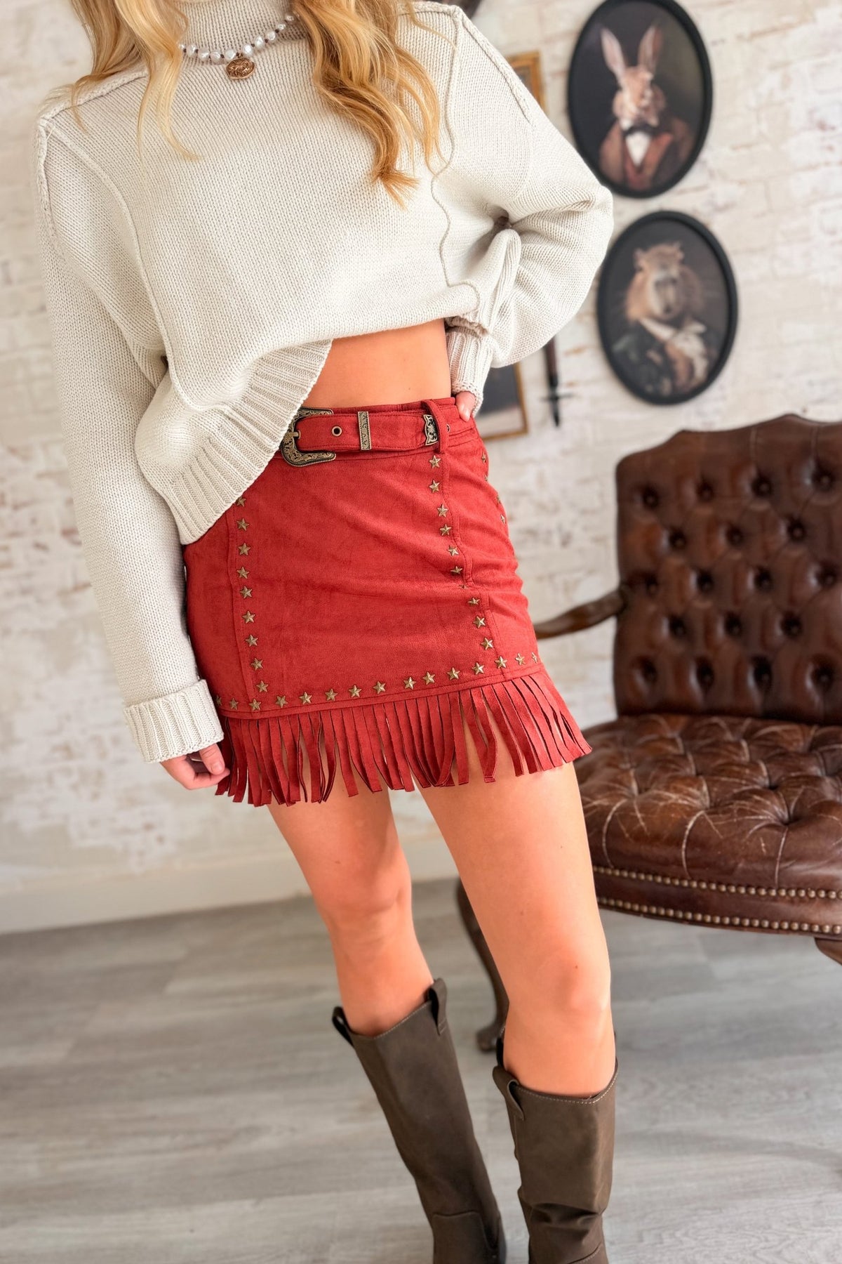 Women’s rust brown suede-look Western mini skirt featuring metallic star stud detailing, a high belted waist with statement cowboy buckle, and long fringe trim along the hem.