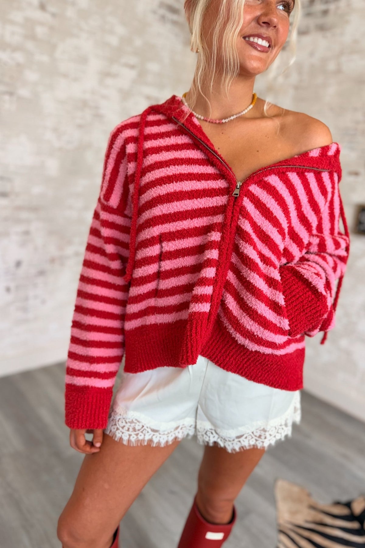 Women’s red and pink striped fuzzy knit zip-up hoodie featuring a soft textured finish, relaxed oversized fit, attached hood with drawstrings, front zipper closure, long sleeves, front pockets, and contrasting red ribbed cuffs and hem.