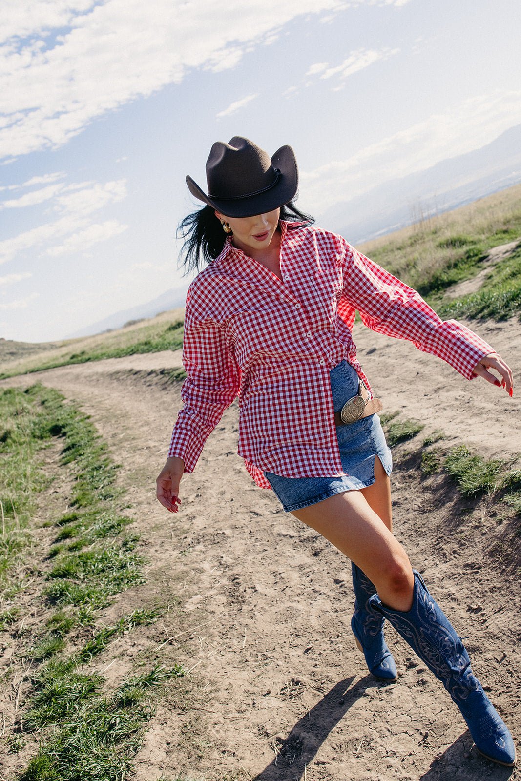 Red and white gingham oversized button-down top styled with a denim mini skirt, western belt, blue cowboy boots, and black cowboy hat for a country concert, rodeo, or Americana-inspired outfit.