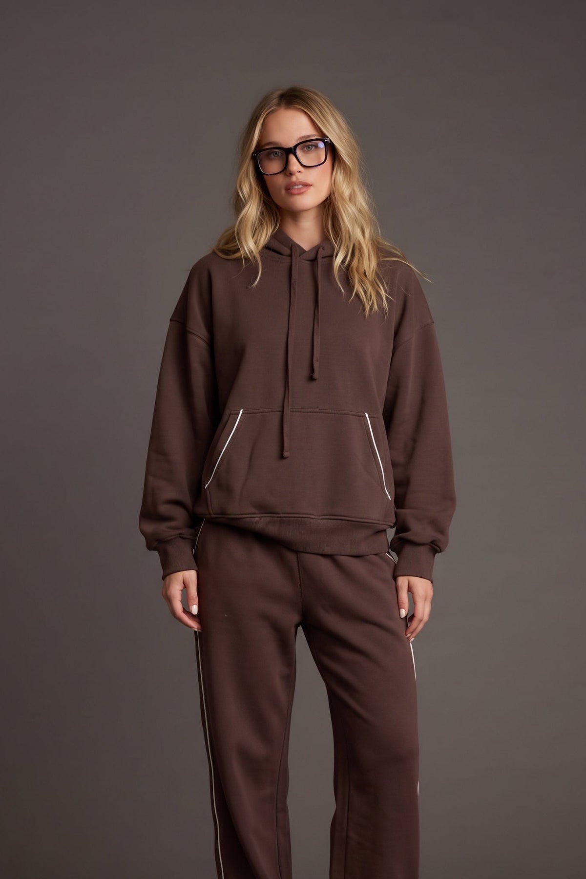 Women’s chocolate brown pullover hoodie featuring a relaxed oversized fit, drawstring hood, dropped shoulders, long sleeves, front kangaroo pocket, and contrast ivory piping detail along the pocket and sides. Coordinating chocolate brown wide-leg sweatpants are available separately for a matching lounge set.