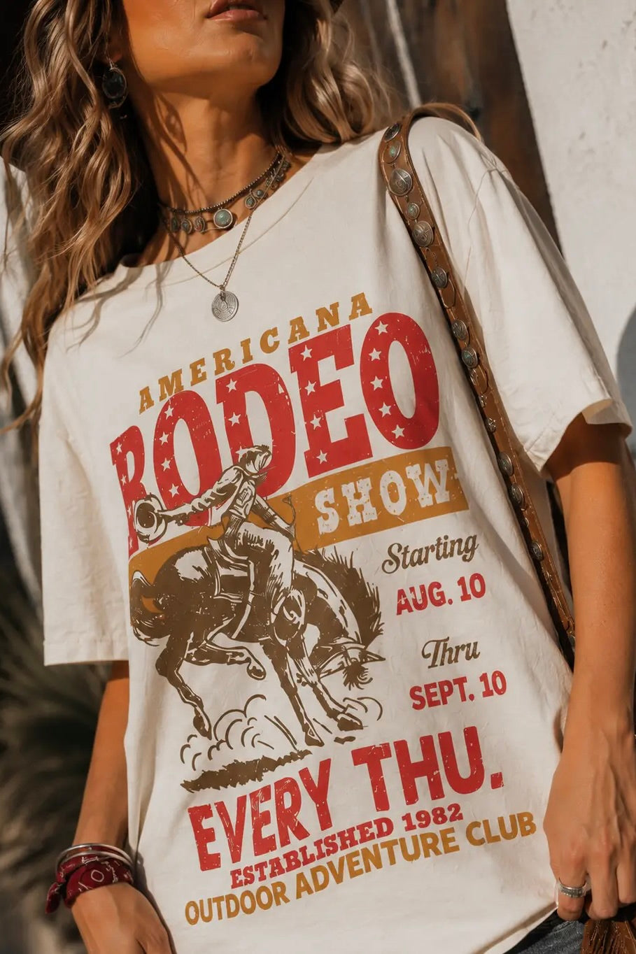 Oversized cream Americana Rodeo graphic tee featuring a vintage bronc rider print with red and gold retro lettering, styled with denim shorts and cowboy boots.